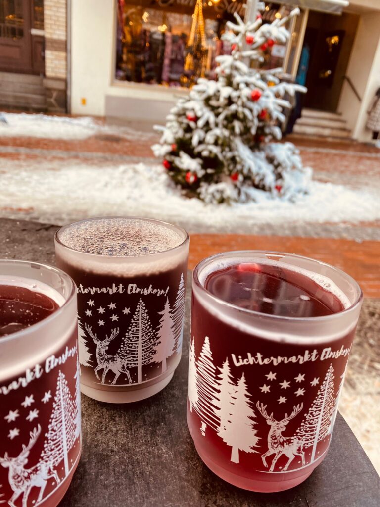 Drei Becher mit Glühwein in weihnachtlichem Ambiente. Auf den Gefäßen steht Lichtermarkt Elmshorn, im Hintergrund ist etwas Schnee auf dem Weihnachtsbaum zu sehen.