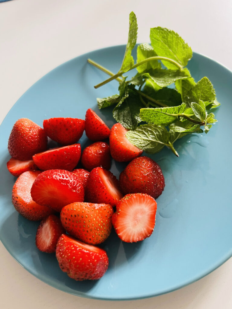 Ein Foto eines hellblauen Tellers, auf dem einige geschnittene Erdbeeren und etwas gezupfte Minze zu sehen sind.