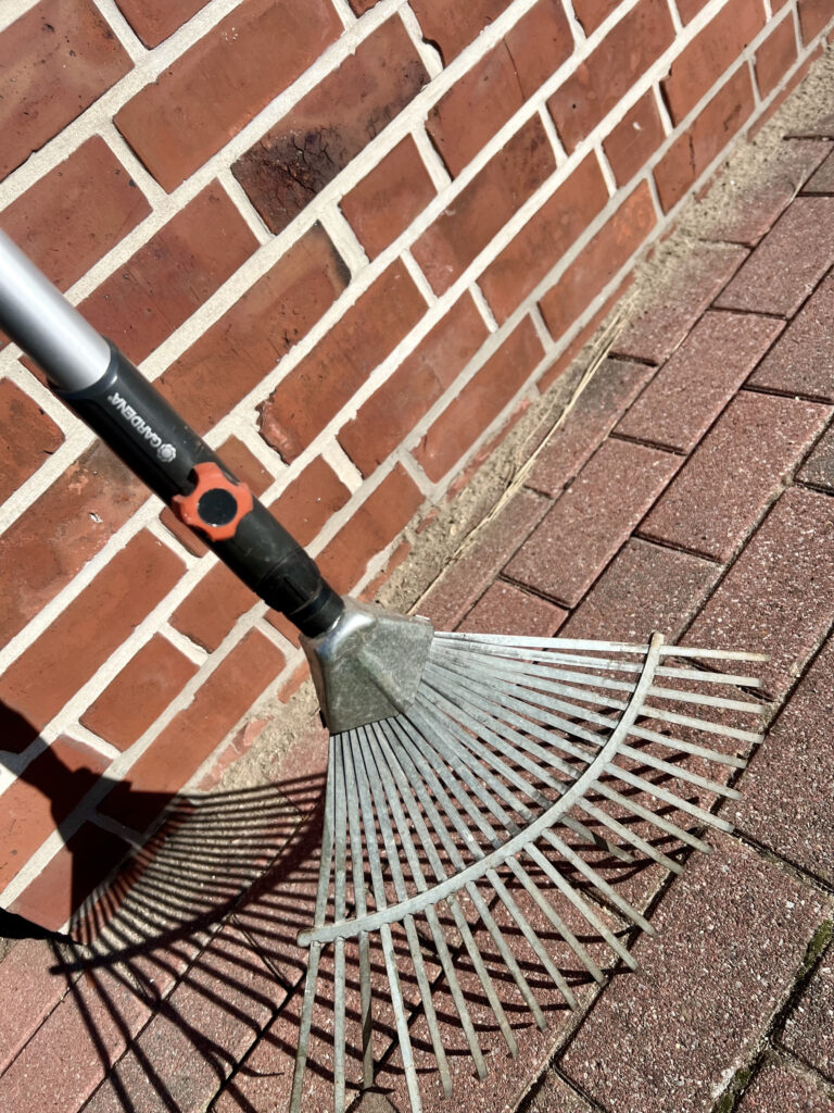 Das untere Ende eines Fächerbesens mit dem breiten Fächer zum Aufnehmen der Blätter. Das Gerät lehnt mit dem Stiel in der Sonne an einer Wand.