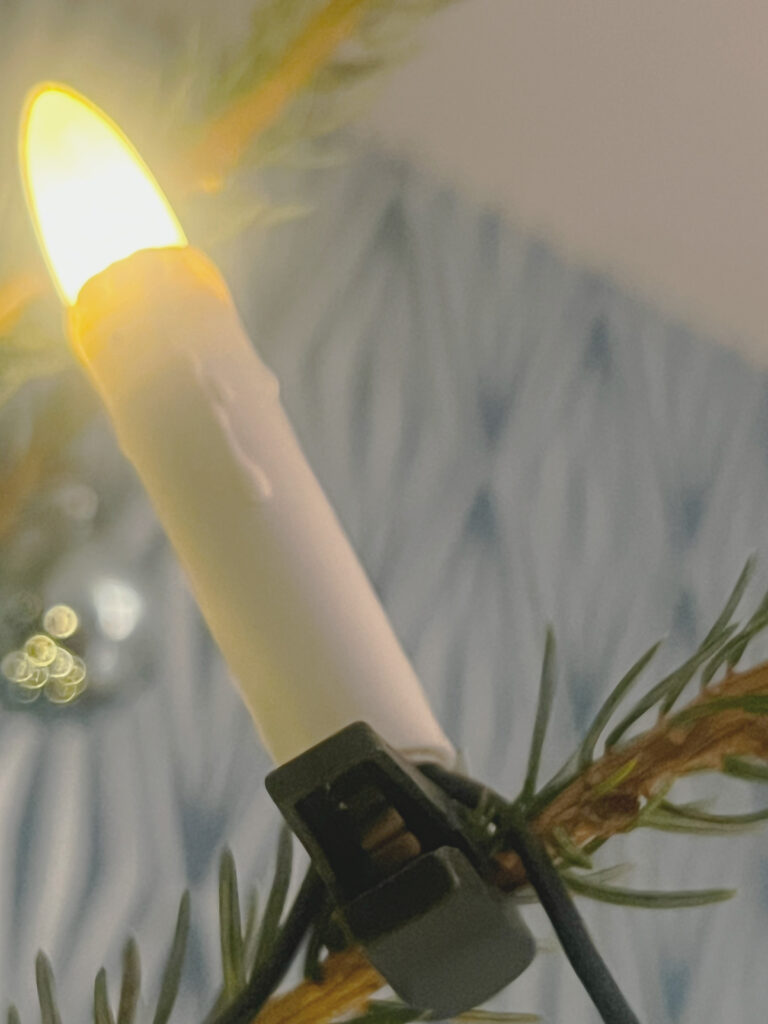 Eine Großaufnahme einer einzelnen Kerze einer LED-Lichterkette am Ast eines Weihnachtsbaum. Sie leuchtet und unten ist der Clip zu sehen, mit dem alles im Baum befestigt wird.