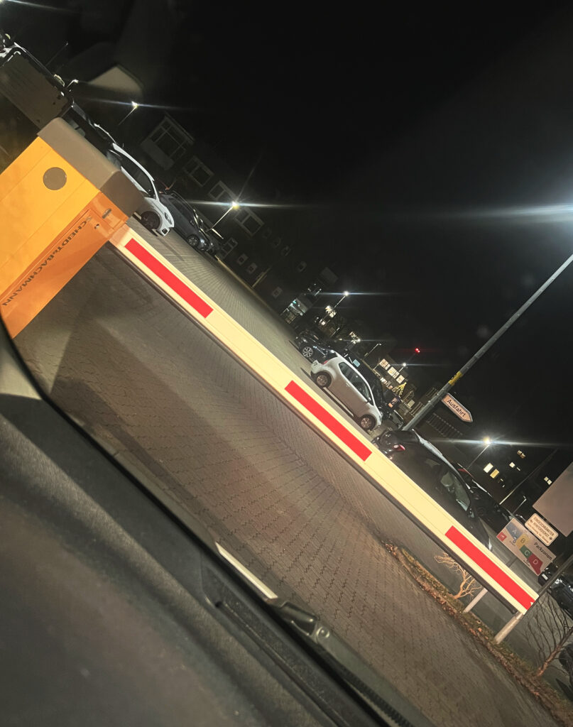 Schräg fotografiert: Eine klassische, moderne Schrankenanlage, die zu einem Parkplatz führt. Die Schranke ist geschlossen, Einlass gibt es nur, wenn links am Automat ein Parkticket gezogen wird.