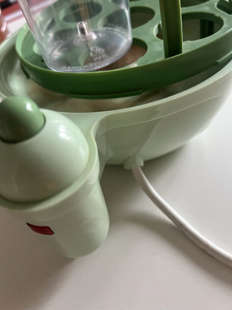 Seitliche Aufnahme eines Eierkochers, der schon ein wenig in die Jahre gekommen ist. Er ist in grünen Farbtönen gehalten, die Heizplatte ist hellgrün, das Gestell für die Eier etwas dunkler. Seitlich ist die Lampe für den Betrieb zu sehen.