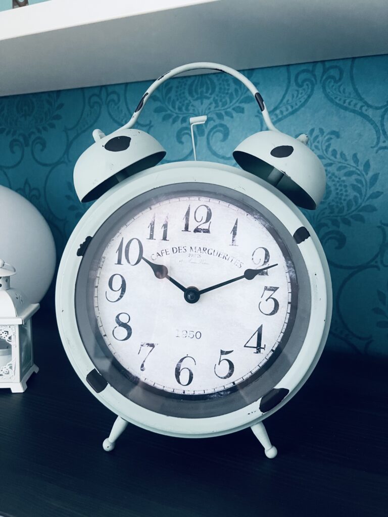 Ein großer Vintage-Wecker vor einer bläulichen Wand, der die Uhrzeit 10:10 Uhr anzeigt und auf der Schriftzug "Café des Marguerites Paris, 1950" zu sehen ist.
