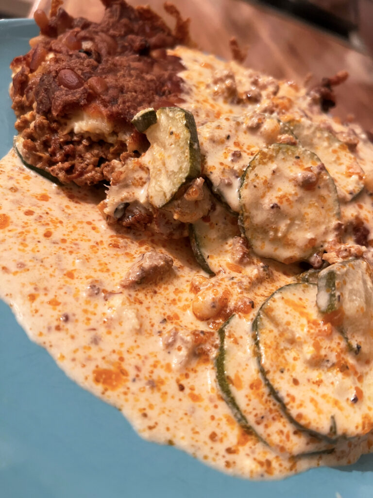 Seitliche Aufnahme eines Tellers mit Zucchini-Hackfleisch-Auflauf mit Feta. Es ist links gut die braune Kruste durch den Käse zu erkennen, auch die dünnen Zucchinischeiben sind gut auszumachen.