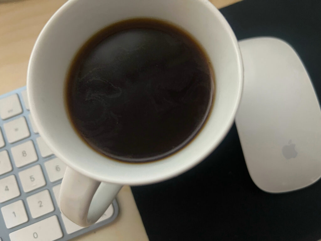 Der Blick von oben in eine Tasse kräftigen schwarzen Kaffee, links davon ist eine Computertastatur zu sehen, rechts davon die Computermaus. Klassische Büroarbeit bei leckerem Duft.