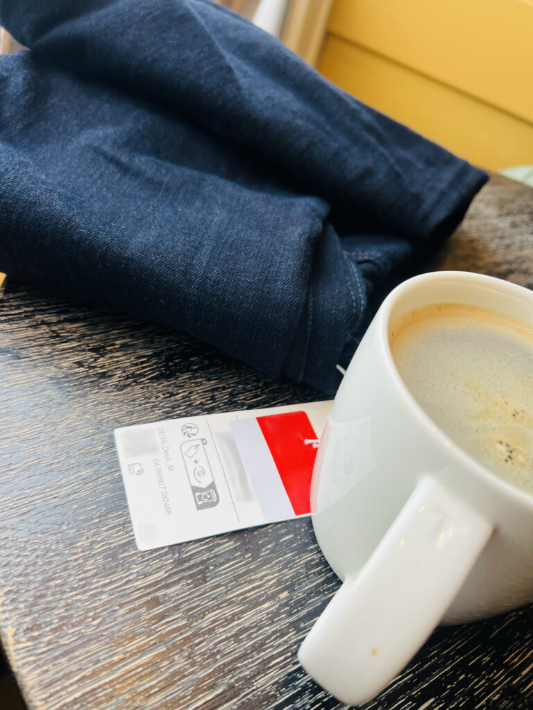 Auf einem Holztisch liegt hinten eine neue Hose, an der vorne noch das Preisschild (rot) zu sehen ist, im Vordergrund steht ein großer Pott Kaffee.